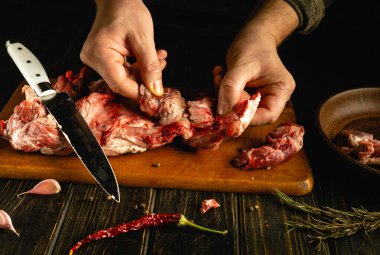 Bir insan otlar, sarımsak ve baharatlarla çevrili ahşap bir tahtada ustalıkla taze et kesip hazırlıyor. Mutfakta sıcak, davetkar bir atmosfer var..