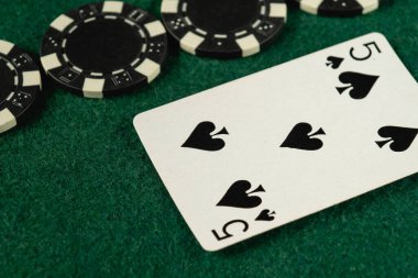 Maça kartlarından beşlisi yeşil keçeli kalemle birlikte birkaç siyah beyaz poker fişi üzerinde duruyor. Bu kurgu, canlı bir kumarhane ortamında poker oynanmasını öneriyor..
