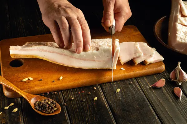 Bir şef, ahşap kesme tahtasıyla domuz göbeğini ustalıkla dilimliyor. Yakınlarda sarımsak ve baharatlar düzenlenerek mutfakta kırsal bir atmosfer yaratılıyor. Taze malzemelerin hazırlanması üzerinde duruluyor..