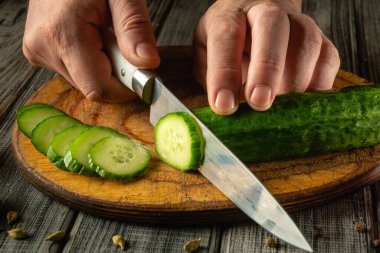 Bir insan taze bir salatalığı tahta bir kesme tahtasının ince dilimlerine ayırıyor. Mutfakta sıcak ve davetkar bir atmosfer var. Malzemelerin tazeliğini vurguluyor..