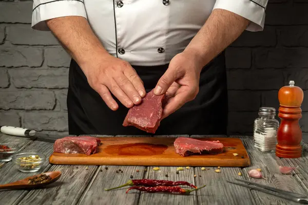 Şef, baharatlar, otlar ve kırsal mutfak zeminiyle çevrili ahşap bir kesme tahtasının üzerinde ustalıkla taze et dilimleri hazırlıyor..