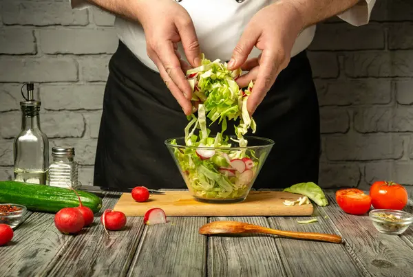 Şefin elleri taze sebzeleri karıştırır, salatalık, marul ve salatalıklar tahta kesme tahtasının üzerinde cam bir kasede..