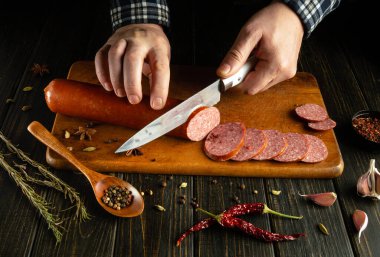 Bir insan, sıcak, davetkar bir mutfak atmosferinde otlar, baharatlar ve taze sarımsakla çevrili ahşap bir kesme tahtasının üzerinde sosisi ustalıkla dilimler..