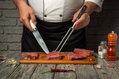 Bir şef, bıçak ve çatal kullanırken ahşap bir tahtanın üzerinde çiğ sığır etini ustalıkla keser. Baharatlar ve baharatlar profesyonel bir mutfakta düzgünce düzenlenir..