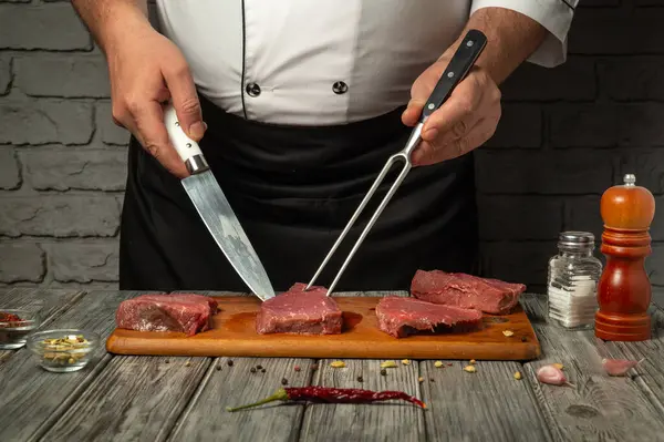 Bir şef, bıçak ve çatal kullanırken ahşap bir tahtanın üzerinde çiğ sığır etini ustalıkla keser. Baharatlar ve baharatlar profesyonel bir mutfakta düzgünce düzenlenir..