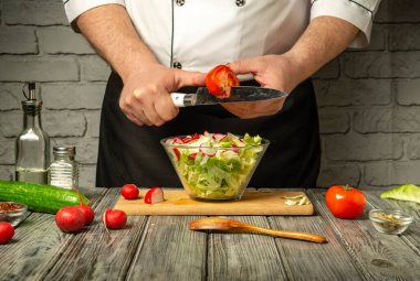 Bir şef, çağdaş bir mutfakta taze bir salata hazırlarken domates dilimliyor. Salatalık ve turp gibi renkli sebzeler şefi çevreleyerek canlı bir atmosfer yaratıyor..