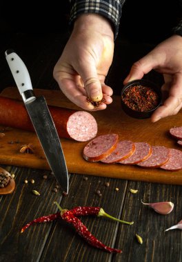Bir elinde taze sarımsak ve biberle çevrili kurutulmuş baharatlar tutarken ahşap bir kesme tahtasının üzerinde füme sosis dilimleyen bir insan. Yakınlarda keskin bir bıçak duruyor..