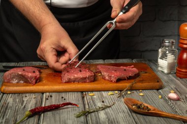 Ahşap bir tahtanın üzerinde eller ustaca sezonlanmış taze sığır eti. Baharatlar ve otlar yakınlarda, et pişirme sanatını ön plana çıkarıyorlar. Aşçının elini çatalla.