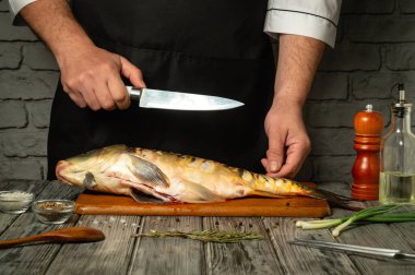 Önlüklü bir şef, ahşap bir tahtanın üzerindeki büyük bir balığı temizlemeye dikkatle odaklanır. Taze otlar, baharatlar ve mutfak gereçleri yakın bir mutfak ortamında düzenlenir..