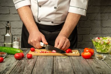 Bir şef ahşap kesme tahtasına domates ve turpları ustaca doğruyor. Taze salatalık ve bir kase karışık salata malzemesi görülebiliyor..