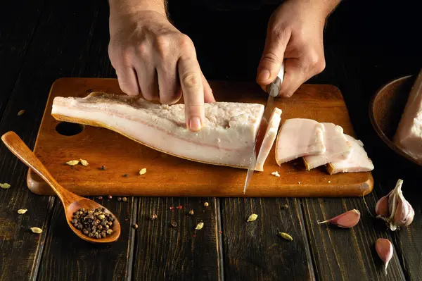 Eller, ahşap bir kesme tahtasındaki kalın bir eti ustalıkla dilimler. Baharat ve sarımsak hazırlığa eşlik ederek, sıcak ve kırsal bir ortamda bir mutfak aracını ön plana çıkarıyor..