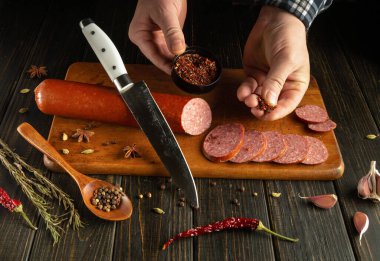 Eller, ahşap kesme tahtasındaki küçük bir kaseye baharat serperken bir sosis kütüğünü dilimliyor. Hazırlık alanını çevreleyen çeşitli otlar ve baharatlar davetkar bir atmosfer yaratıyor..