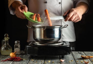 Aşçı ocağın tepesindeki tencereye sosisli sandviç ekler, yemek malzemeleriyle çevrili. Renkli baharatlar mutfak atmosferini geliştirirken buhar yükselir..