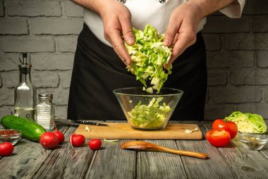 Yetenekli bir şef sebze doğruyor, marul ve domates de dahil, ve cam bir kasede karıştırıyor. Taze malzemeler ve mutfak aletleri ahşap bir tezgaha saçılmış..