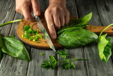 Elleri ustalıkla taze ıspanak ve otları tahta kesme tahtasında dilimliyor. Kırsal mutfak ortamında doğal ışık, canlı yeşil renkler ve organik dokular vurgulanıyor.