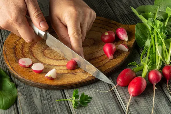 Kırsal ahşap bir kesme tahtasındaki canlı kırmızı turpları ustalıkla dilimler. Yakınlarda bir sürü taze yeşillik görünüyor. Bu da yemek hazırlığını gösteriyor..