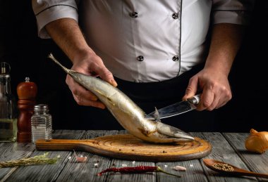 Bir şef, sıcak bir mutfakta taze balık keserken uzman tekniği gösterir. Ahşap kesim tahtasının etrafını saran çeşitli malzemeler ve çatal bıçaklar aşçılık yaratıcılığıyla dolu bir akşamı vurguluyor..