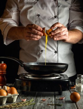 Beyaz üniformalı bir şef, fırında tavanın üstünde yumurta kırıyor. Taze yumurta ve bitkiler ahşap bir masaya dizilir ve sıcak bir yemek atmosferi yaratır..