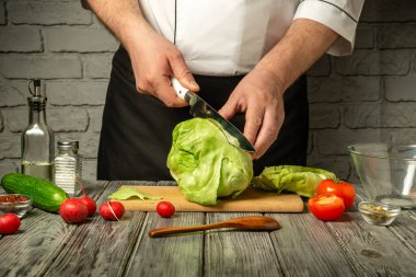 Bir şef ahşap kesme tahtasıyla lahana kafasını ustalıkla dilimliyor. Etrafını saran malzemeler arasında domates, salatalık ve turplar var..
