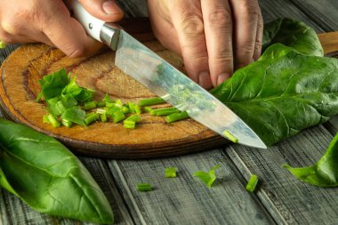 Eller ahşap bir kesim tahtasının üzerinde ustalıkla taze ıspanak doğruyor, rahat bir mutfakta vitamin zengini bir yemek hazırlıyor. Güneş ışığı sebzelerin parlak rengini artırır..