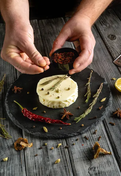 İki el kurutulmuş kırmızı biber ve otları siyah bir tabağa yerleştirilmiş yuvarlak peynir diskine serpiyor..