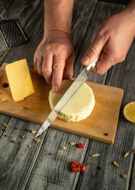Eller, ahşap bir kesme tahtasındaki yuvarlak peyniri ustalıkla dilimler. Etrafını saran baharatlar ve limon mutfak ortamını daha da güzelleştiriyor..