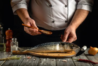 Bir şef taze uskumru balığını baharat serpmek için ahşap bir kaşıkla çeşniliyor. Kırsal mutfak atmosferinde tuz, biber ve soğan gibi malzemeler bulunur..