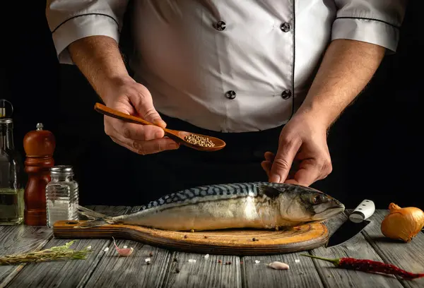 Bir şef taze uskumru balığını baharat serpmek için ahşap bir kaşıkla çeşniliyor. Kırsal mutfak atmosferinde tuz, biber ve soğan gibi malzemeler bulunur..