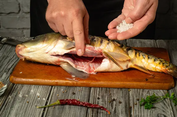 Eller taze bir balığı dikkatlice temizliyor, pişirirken tuz ve baharat ekliyor. Mutfakta kırsal ahşap bir tahta ve canlı malzemeler var..
