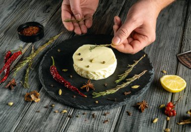 Eller, yuvarlak bir peyniri çeşitli aromalı malzemelerle çevrili koyu bir tahta üzerinde taze otlar ve baharatlarla süslüyor..