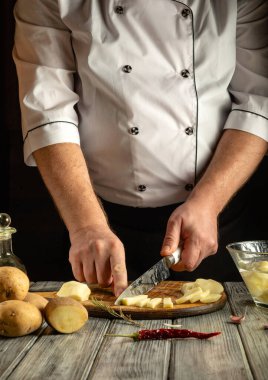 Beyaz üniformalı bir şef ahşap bir kesim tahtasının üzerinde patates ve kırmızı biber doğruyor. Sıcak ışık sıcacık mutfak atmosferini güçlendiriyor..