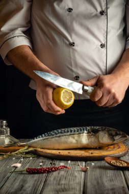 Bir şef, ahşap bir tahtanın üzerinde bütün bir balığın üzerinde bıçak ve yarım limon tutar. Çeşitli malzemeler ve malzemeler etrafa saçılmış, sıcak ve rahat bir atmosfer oluşturuyorlar..