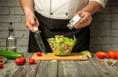 Bir şef ustalıkla taze sebzeleri doğrar ve bir kase salataya tuz ekler. Mutfakta ahşap yüzeyli kırsal bir dekor var. Sıcak bir atmosfer yaratıyor..