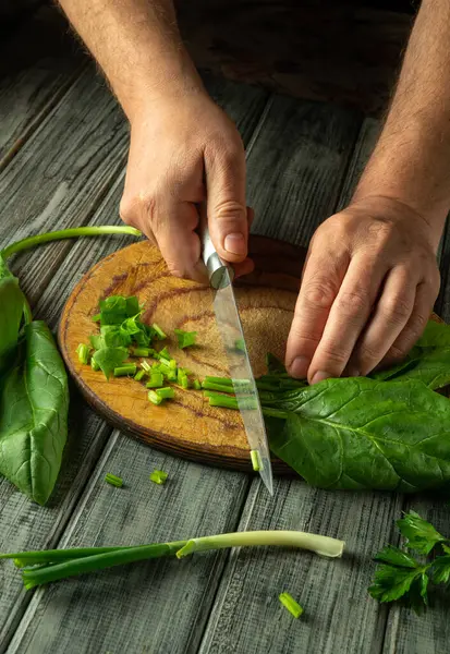Ellerin ustalıkla yeşil soğanları ve yapraklı yeşillikleri ahşap bir kesme tahtasının üzerinde doğruyor, rahat bir mutfakta taze yemek hazırlığının özünü yakalıyor..