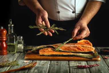 Bir şef ustalıkla taze somonu otlarla süslüyor, ahşap tezgahın başında temel yemek malzemeleriyle duruyor. Ilık aydınlatma mutfak atmosferini güçlendiriyor..