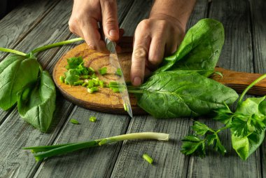 Bir insan taze ıspanak ve yeşil otları kesmek için tahta kesme tahtasına keskin bir bıçak kullanır. Mutfak seti ahşap dokuları olan bir köy atmosferi sergiliyor..
