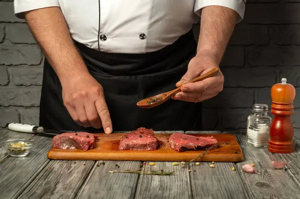 Bir şef ahşap kesme tahtasındaki üç parça eti çeşnilendirmeye odaklanır. Taze baharatlar ve baharatlar yakınlarda, kırsal bir mutfak ortamındaki aşçılık sürecini vurguluyorlar..
