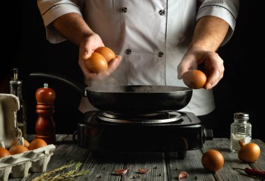 Loş bir mutfakta, bir aşçı tavanın üstünde iki yumurta tutar ve birkaç yumurta etrafta otururken onları pişirme sürecine eklemeye hazırdır..