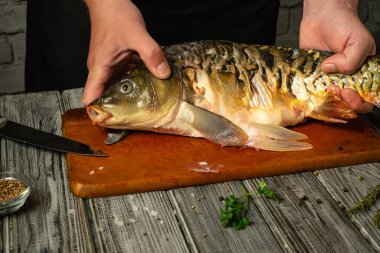 Bir insanın elleri, filetoya hazırlanırken tatlı su balığını ahşap bir kesme tahtasının üzerinde tutar. Yakınlarda çeşitli otlar ve baharatlar görülebiliyor. Mutfak atmosferi yaratıyorlar..