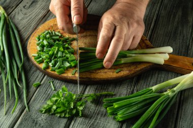 Elleri ustalıkla taze soğan ve otları doğramak ahşap bir kesim tahtasına, çevresine yayılmış canlı yeşillikler. Doğal ışık, bu mutfaktaki taze malzemelerin cazibesini arttırır..