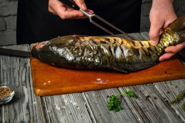 Tahta kesme tahtasındaki bir balığı hazırlamak için çatal ve bıçağı ustalıkla kullanan bir insan. Taze bitkiler ve baharatlar mutfağın canlı atmosferine eklenir..