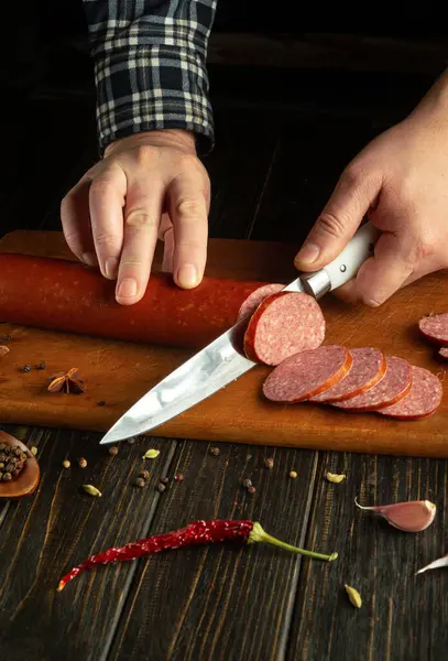 Bir el, baharatlı sosisi ahşap kesme tahtasındaki mermiler halinde ustalıkla dilimler. Çeşitli baharatlar ve tüm biberler tahtanın etrafını sararak, kırsal mutfak atmosferine katkı sağlıyor..