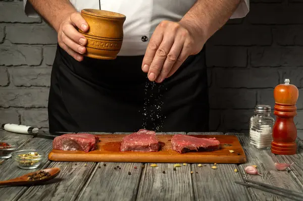 Bir şef, ahşap bir kesim tahtasındaki çiğ biftekleri ustalıkla mevsimlendirir. Etrafı çeşitli otlar ve baharatlarla çevrilidir..