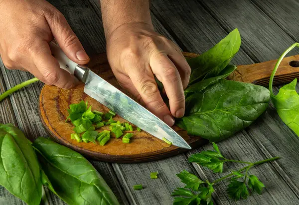 Elleri ahşap bir tahtanın üzerinde taze sebze ve sebzeleri ustalıkla kesiyor. Yanında canlı yapraklar ve doğranmış sebzeler var..