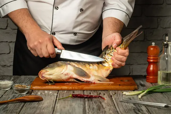 Bir şef, bir balığı ahşap kesme tahtasına saplamaya hazır bir şekilde ustalıkla tutar. Mutfakta, yanında baharatlar ve yeşil soğanlar olan kırsal bir ortam var. Taze sazan temizliyorum..