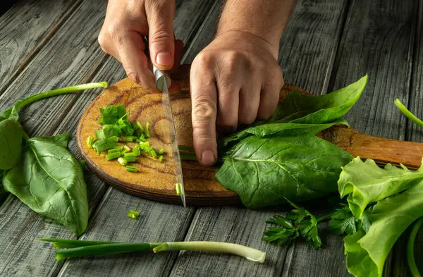 Doğal ışığın ortasında, rahat bir mutfakta, taze yeşilliklerle çevrili ahşap bir tahtanın üzerinde, ustaca yeşil soğan ve ıspanak kesen eller..