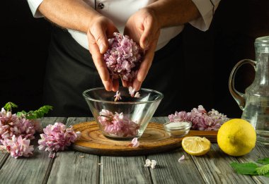 Bir şef ustalıkla akasya çiçeklerini limon dilimleriyle karıştırır. Taze bitkiler ve su, ahşap bir masada canlı malzemelere eşlik ederek, mis kokulu bir karışım oluşturur..
