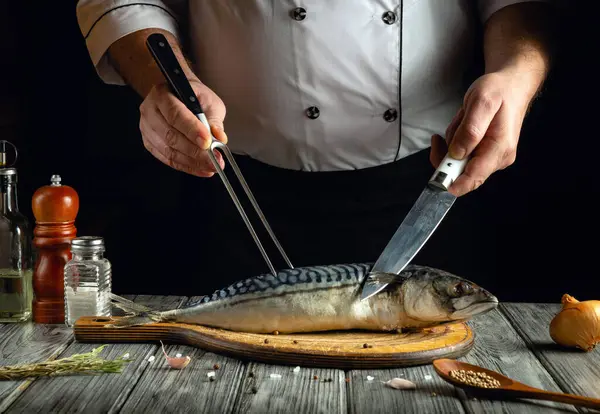 Bir şef, ahşap bir kesme tahtasındaki uskumruyu ustalıkla fileto yapıyor. Bıçak ve çatal kullanıyor. Etrafı baharat ve mutfak aletleriyle çevrili..