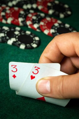 Poker meraklısı, yeşil keçeli masa etrafına çeşitli renkli çipler saçılırken, bir elinde iki üçlü tutarak oyunun kritik bir anına hazırlanıyor..