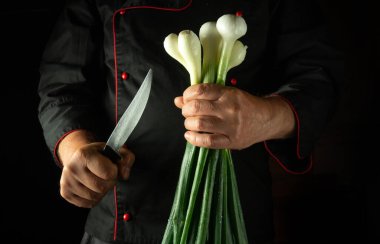Bir şef loş bir mutfakta duruyor, bir elinde taze soğanları tutarken diğer elinde keskin bir bıçak, doğramaya hazır..